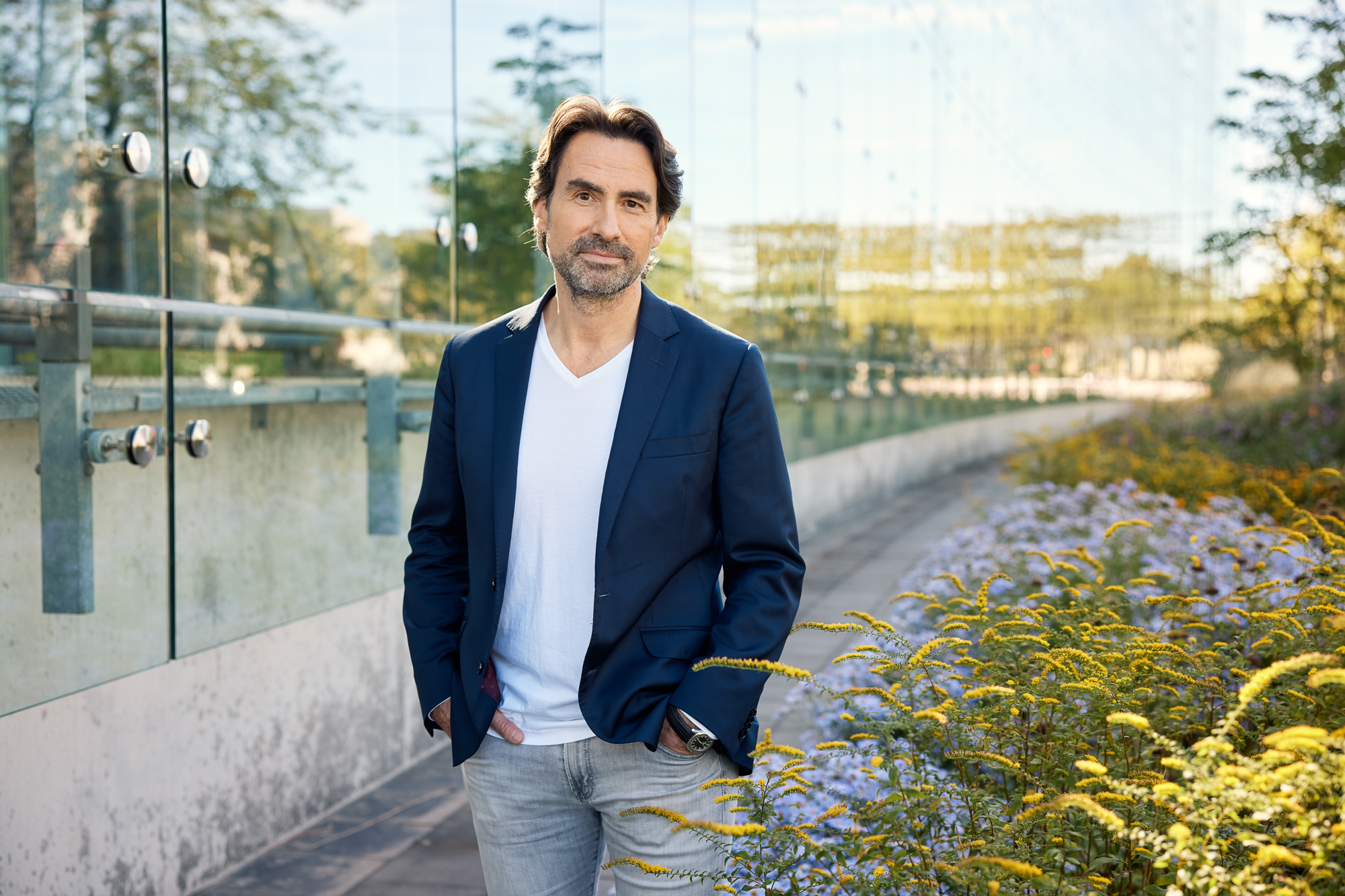 Ein Mann mit dunklem Haar und Bart steht im Freien und sieht aus wie ein Reportagefotograf aus Düsseldorf. Er trägt einen marineblauen Blazer und ein weißes Hemd auf einem mit Glasgeländern und gelben Blumen geschmückten Weg. Portrait und Reportage Fotografie für Corporate und Editorial
