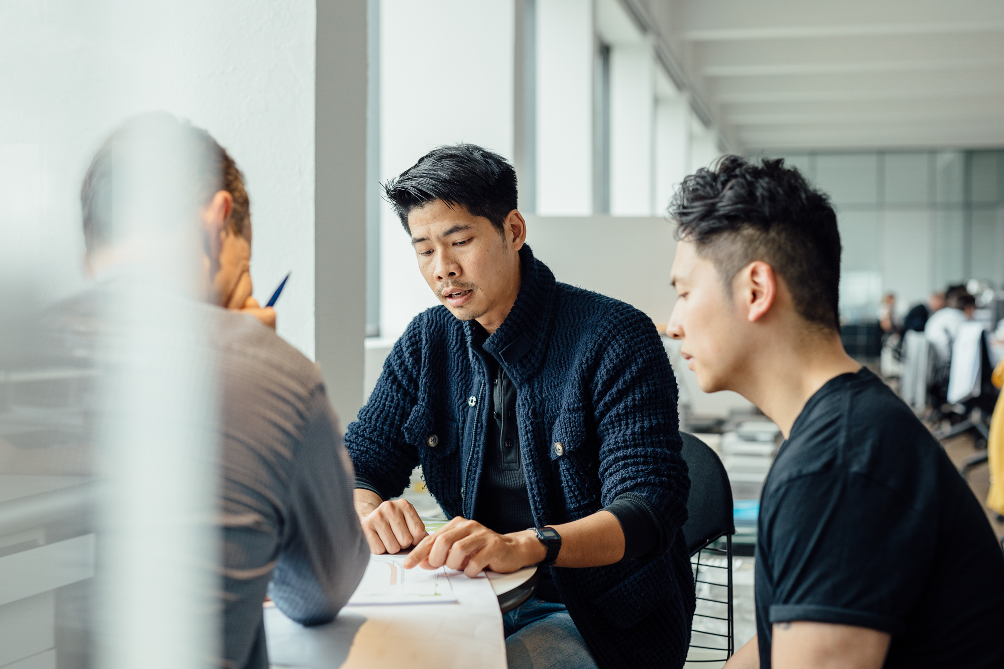 In einem Düsseldorfer Büro sitzen drei Personen an einem Tisch und diskutieren intensiv über Employer Branding. Eine Person gestikuliert leidenschaftlich, während die anderen aufmerksam zuhören. Ihre Ausdrücke ähneln professionellen Porträts und fangen die Essenz der Zusammenarbeit ein. Portrait und Reportage Fotografie für Corporate und Editorial