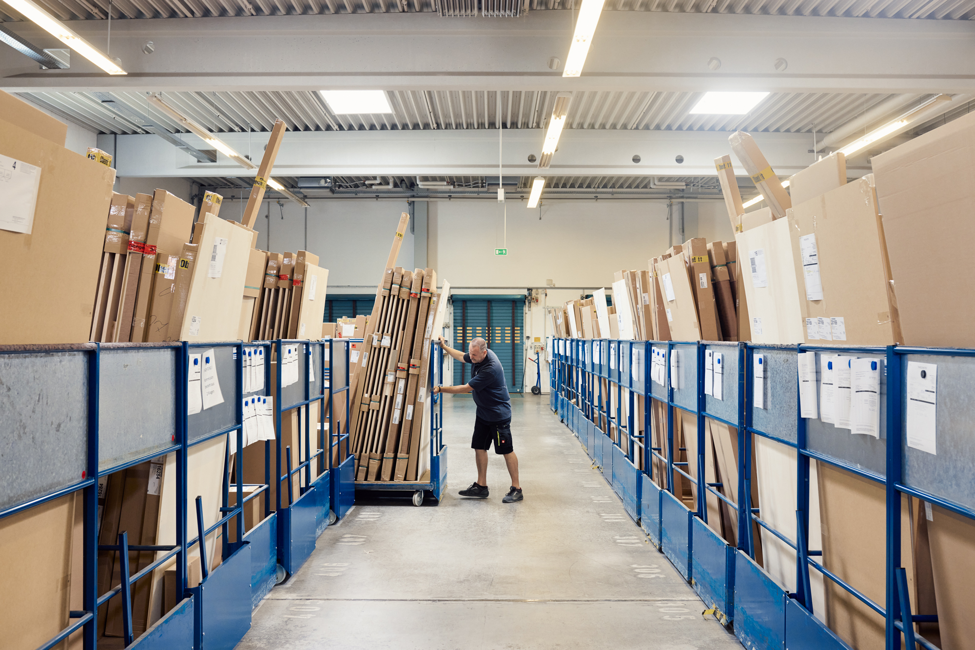 In einem geschäftigen Düsseldorfer Lager manövriert ein Arbeiter einen mit großen Kartonpaketen beladenen Wagen durch einen Gang mit ähnlichen Waren und fängt so die typische Hektik der Unternehmenslogistik ein. Portrait und Reportage Fotografie für Corporate und Editorial