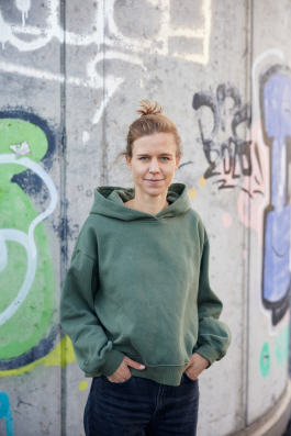Eine Person in einem grünen Kapuzenpulli steht vor einer mit Graffiti beschmierten Betonwand und blickt mit den Händen in den Taschen in die Kamera, was eine urbane Variante des Personal Branding darstellt. Portrait und Reportage Fotografie für Corporate und Editorial