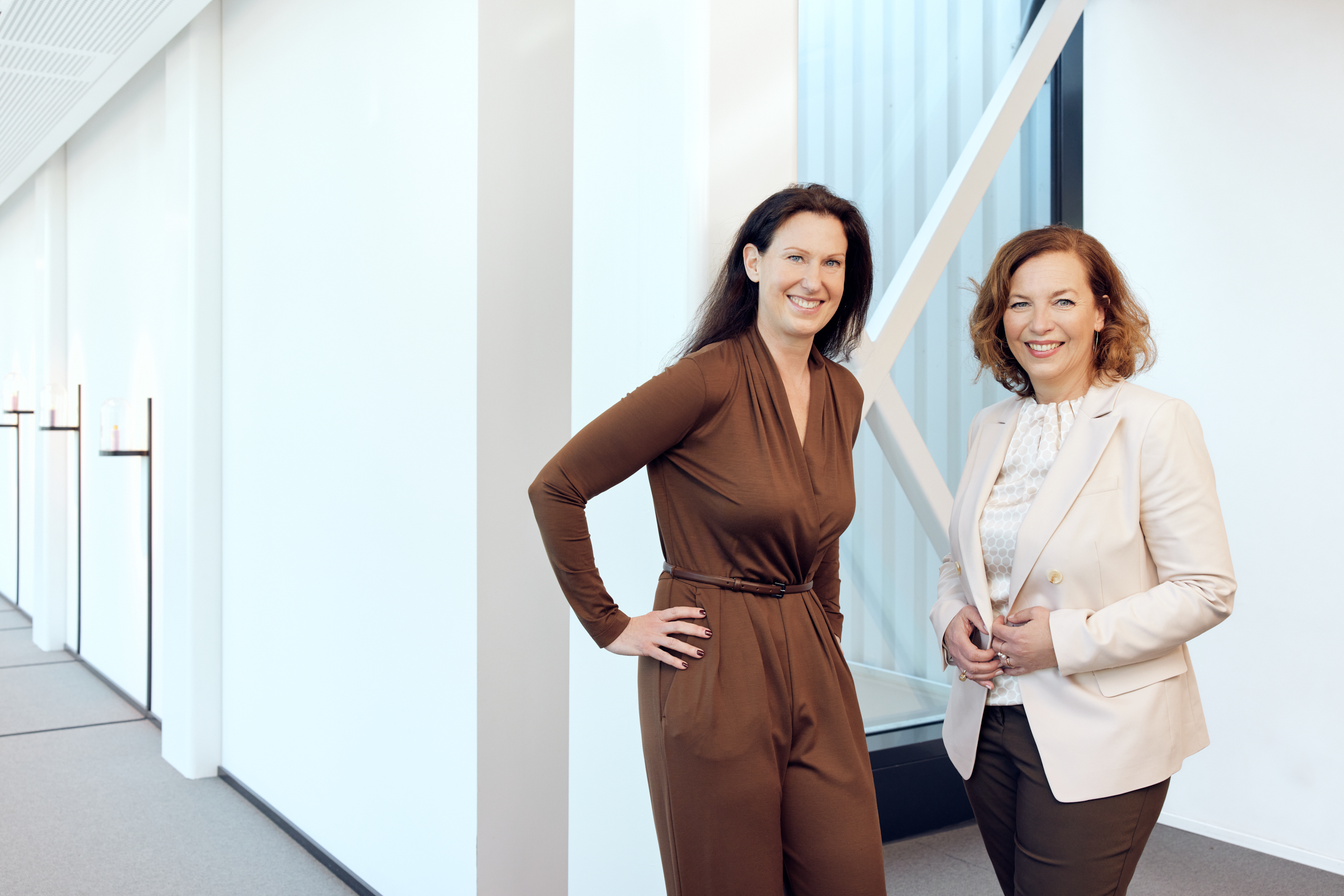 Zwei Frauen stehen in einem modernen, gut beleuchteten Flur und lächeln herzlich zum Mitarbeitershooting. Die eine trägt ein braunes Kleid, die andere ein beiges Kostüm mit weißer Bluse, was ein natürlich einladendes Gruppenbild ergibt. Portrait und Reportage Fotografie für Corporate und Editorial