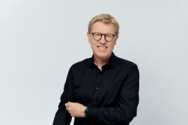 Eine Person mit Brille und schwarzem Hemd lächelt vor einem schlichten Hintergrund in die Kamera und fängt die Essenz eines professionellen Businessporträts ein. Portrait und Reportage Fotografie für Corporate und Editorial