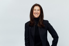 Eine lächelnde Frau mit langen braunen Haaren, gekleidet in einen schwarzen Blazer und Rollkragenpullover, steht vor einem schlichten weißen Hintergrund und fängt die Essenz eines professionellen Businessporträts ein, das ideal für Mitarbeiter in NRW ist. Portrait und Reportage Fotografie für Corporate und Editorial