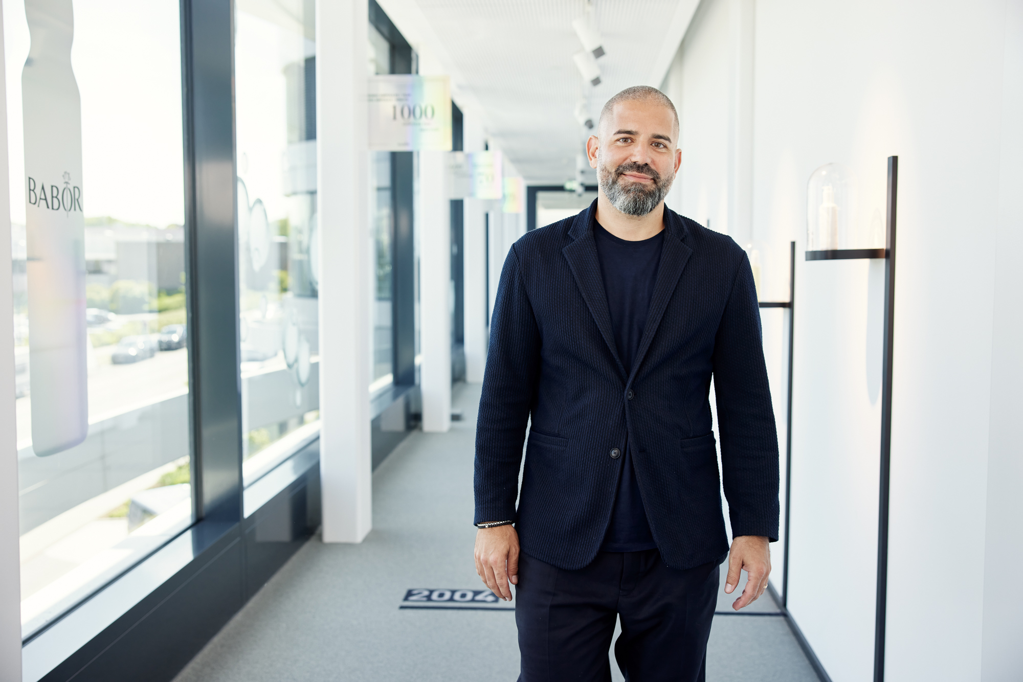 Ein Mann im marineblauen Anzug und schwarzem T-Shirt steht authentisch in einem hellen Flur mit großen Fenstern und bietet einen porträtartigen Blick nach draußen, der an einen Geschäftsführer mit Führungsverantwortung erinnert. Portrait und Reportage Fotografie für Corporate und Editorial