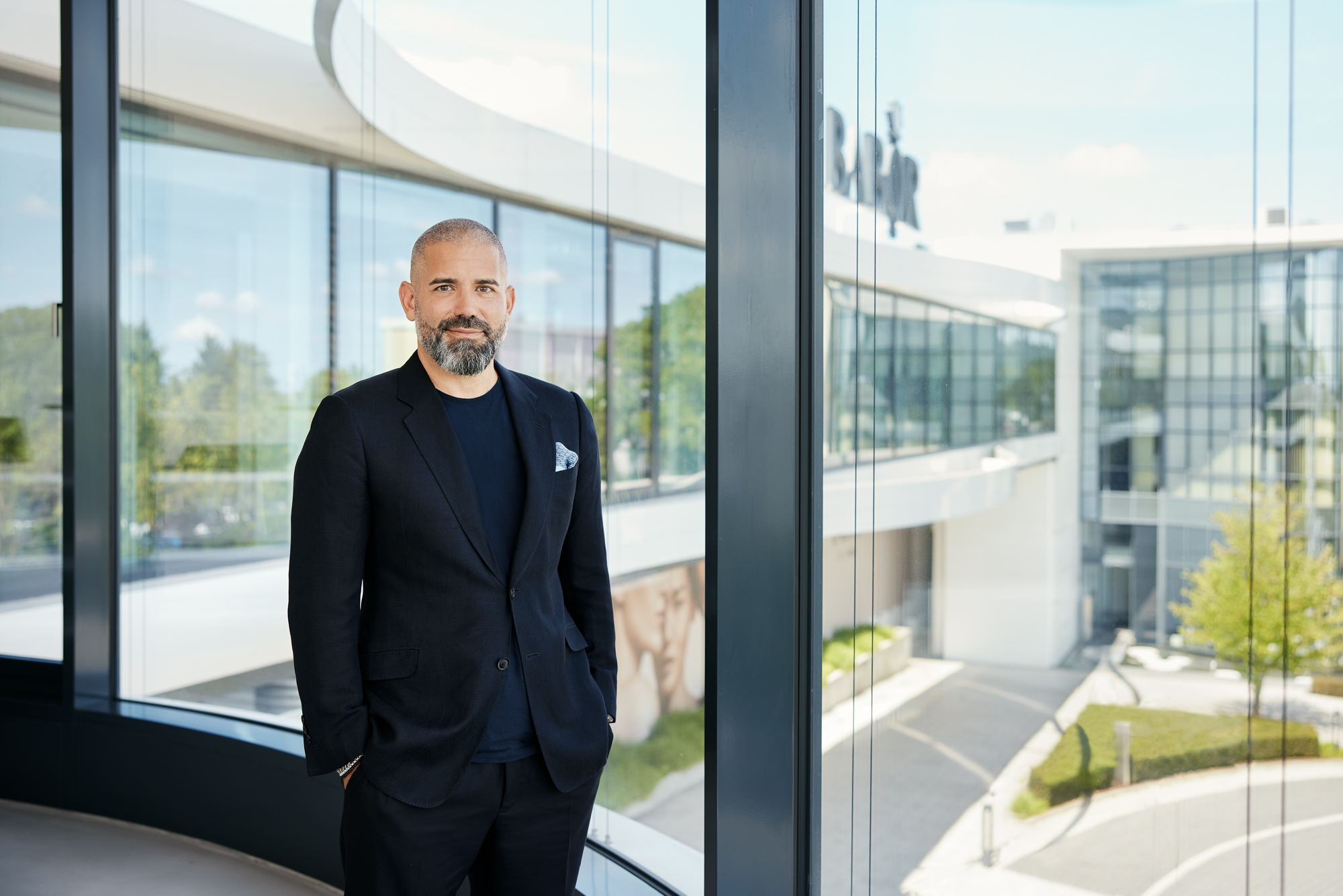 Ein Geschäftsführer im schwarzen Anzug steht neben großen Fenstern. Die schlichte Architektur eines modernen Gebäudes bietet die perfekte Kulisse für dieses professionelle Porträt in NRW. Portrait und Reportage Fotografie für Corporate und Editorial