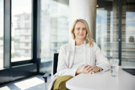 Auf einem eleganten Businessportrait sitzt eine Person mit langen Haaren an einem Schreibtisch in Aachen, die Hände gefaltet und in einen hellen Blazer gehüllt. Auf dem Tisch steht ein Glas Wasser, was auf einen Moment der Besinnung inmitten der Management-Pflichten hindeutet. Portrait und Reportage Fotografie für Corporate und Editorial
