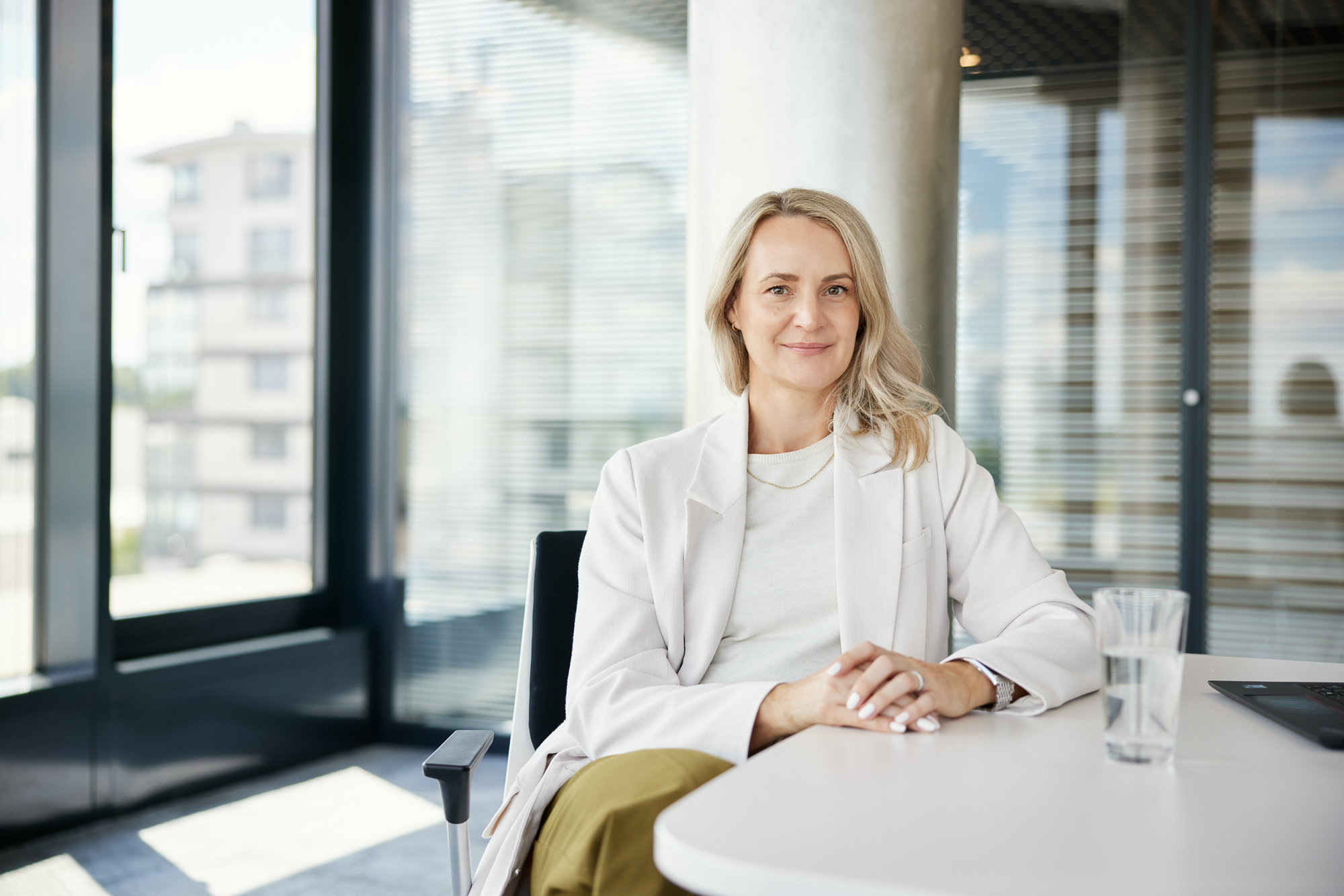 Auf einem eleganten Businessportrait sitzt eine Person mit langen Haaren an einem Schreibtisch in Aachen, die Hände gefaltet und in einen hellen Blazer gehüllt. Auf dem Tisch steht ein Glas Wasser, was auf einen Moment der Besinnung inmitten der Management-Pflichten hindeutet. Portrait und Reportage Fotografie für Corporate und Editorial