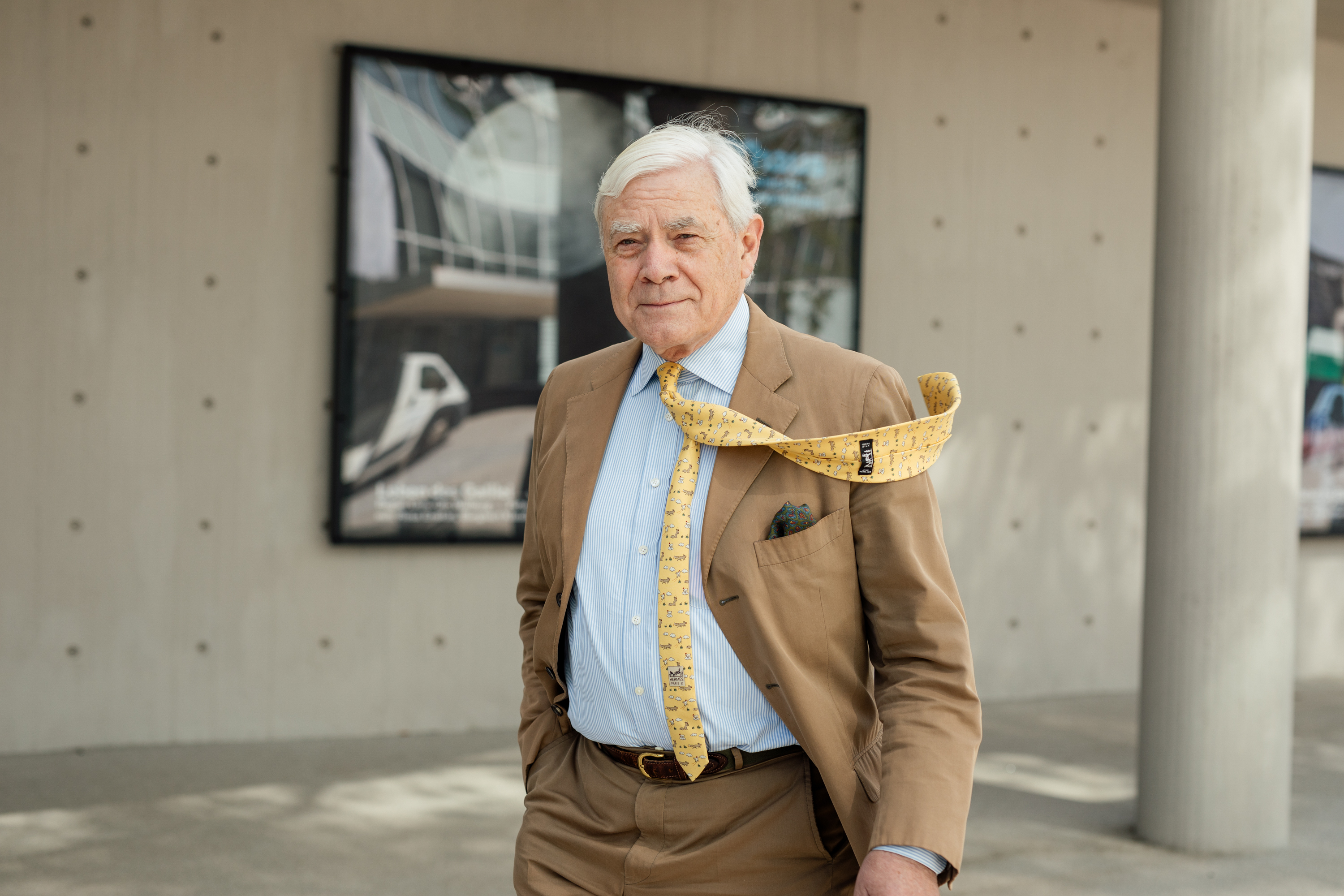 Ein älterer Mann im hellbraunen Anzug und mit gelber Krawatte, der vom Wind zur Seite geweht wird, steht im Freien vor einem modernen Gebäude mit Plakaten - ein eindrucksvolles Geschäftsporträt, das die persönliche Markenbildung in einem Unternehmensumfeld festhält. Portrait und Reportage Fotografie für Corporate und Editorial