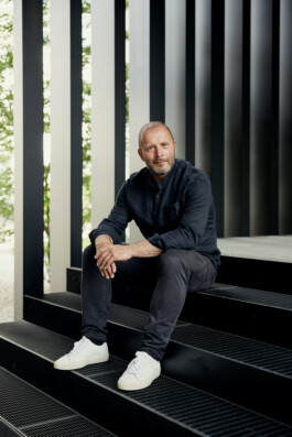 Auf schwarzen Metallstufen sitzt eine Person mit Bart. Sie trägt ein dunkles Hemd, dunkle Hosen und weiße Turnschuhe. Im Hintergrund sind vertikale Lamellen und ein Hauch von Grün zu sehen. Portrait und Reportage Fotografie für Corporate und Editorial