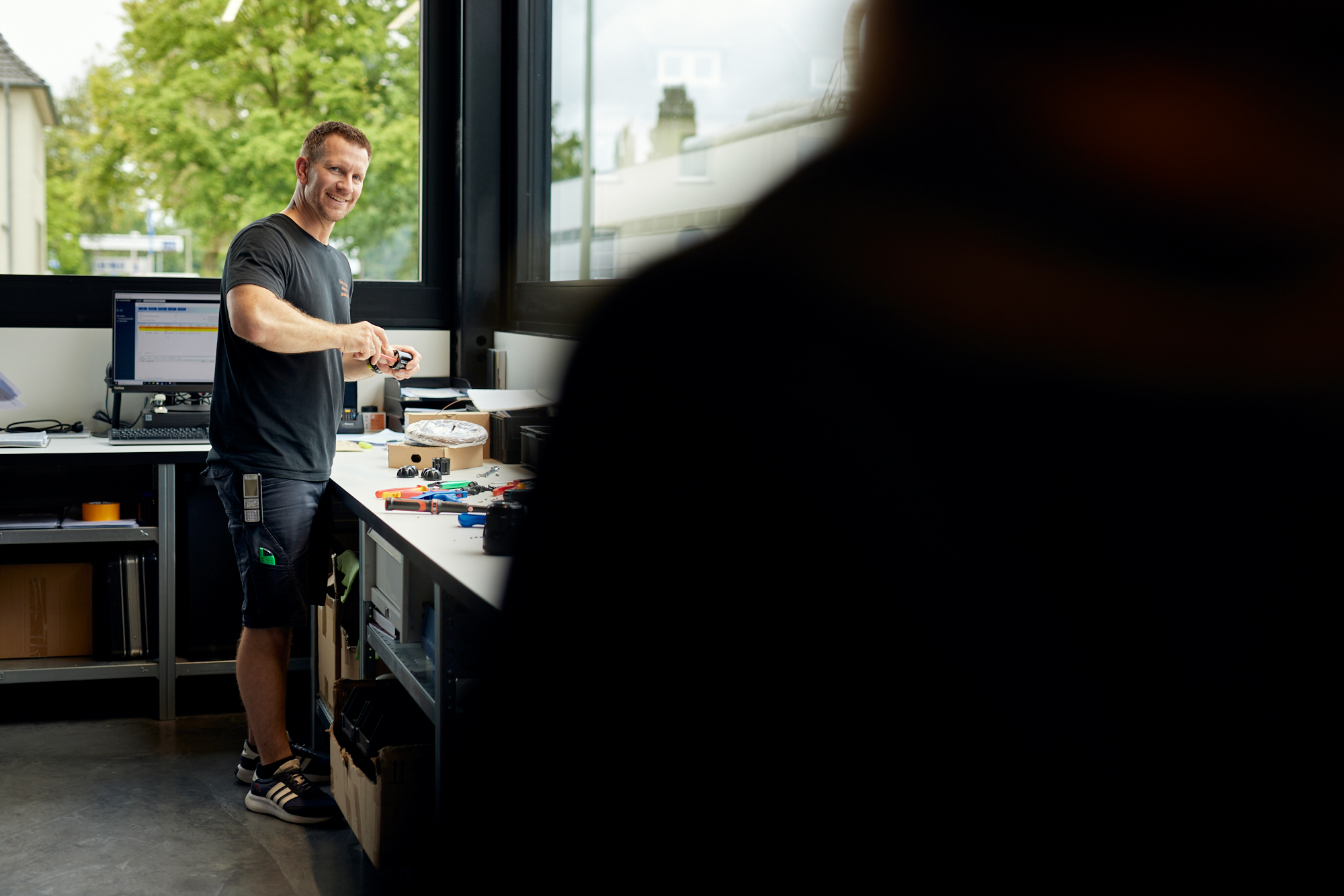 Ein Mann lächelt in einer Werkstatt über einer mit Werkzeugen gefüllten Werkbank, während die Silhouette einer anderen Person diesem Imagefilm Spannung verleiht und sich perfekt eignet, um in den sozialen Medien Aufmerksamkeit zu erregen. Portrait und Reportage Fotografie für Corporate und Editorial