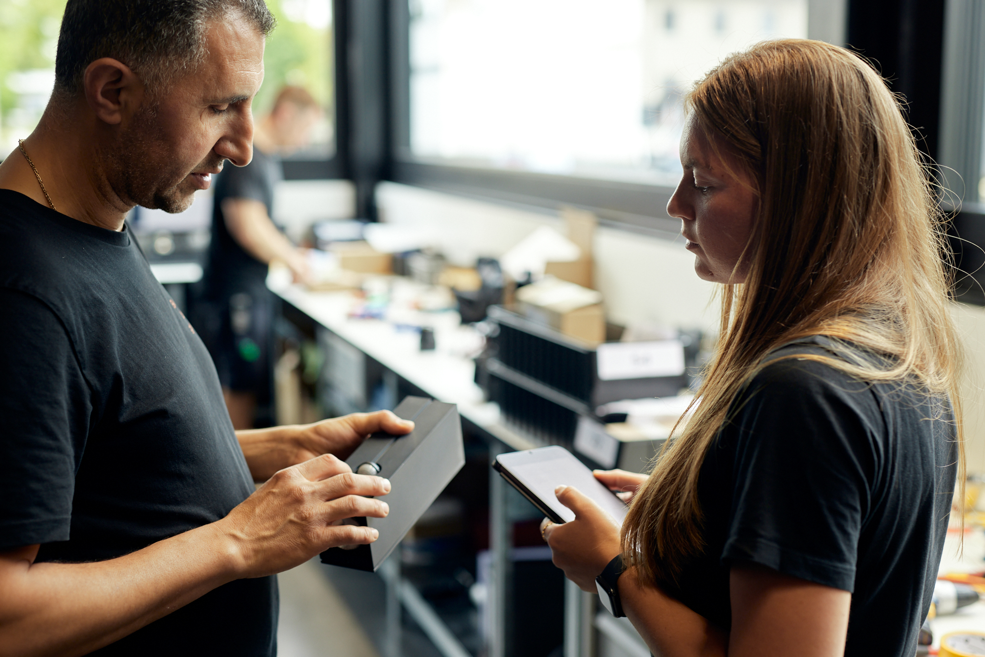 Zwei Personen stehen im Innenbereich und diskutieren über Gegenstände in ihren Händen. Einer hält einen rechteckigen Gegenstand, während der andere ein Tablet betrachtet. Im Hintergrund sind Regale mit verschiedenen Gegenständen zu sehen – perfekt für einen Imagefilm oder Video-Content für Ihre Website und Social Media-Plattformen. Portrait und Reportage Fotografie für Corporate und Editorial