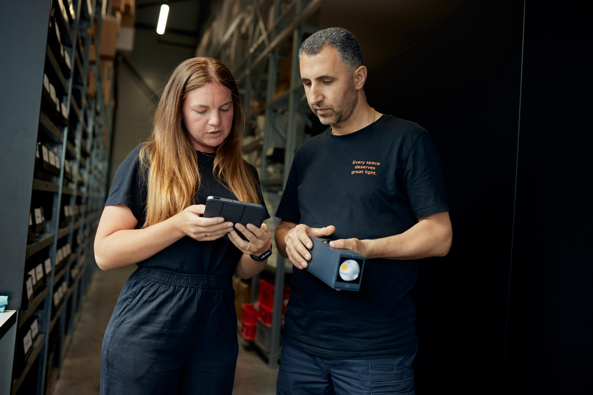 In einem Lagerraum diskutieren zwei Personen in schwarzen Hemden über ein Gerät – eine mit einem Tablet, die andere mit einem kleinen elektronischen Gerät. Regale mit Kisten füllen den Hintergrund und bilden eine ideale Kulisse für dynamische Imagefilme oder die Erstellung ansprechender Foto- und Videoinhalte. Portrait und Reportage Fotografie für Corporate und Editorial