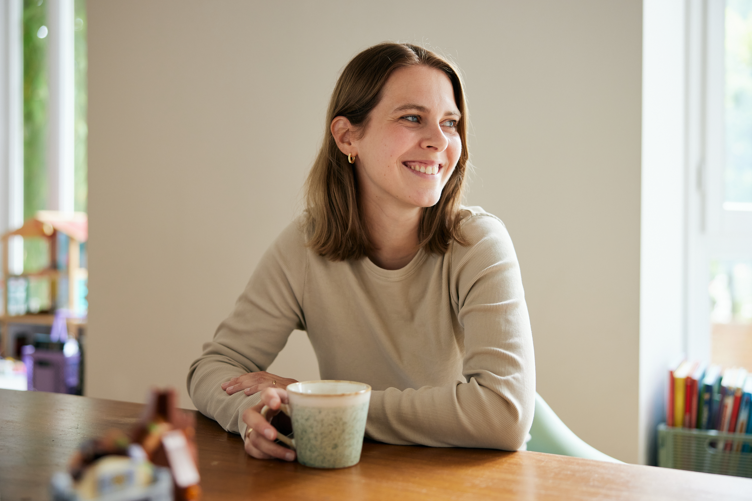 Eine Frau mit schulterlangen braunen Haaren sitzt an einem Holztisch, lächelt und hält eine gesprenkelte Tasse in der Hand - ein warmes und sympathisches Business-Porträt, das ihre Persönlichkeit einfängt und sich perfekt für das Personal Branding eignet. Bücher und ein Fenster füllen den Hintergrund. Portrait und Reportage Fotografie für Corporate und Editorial