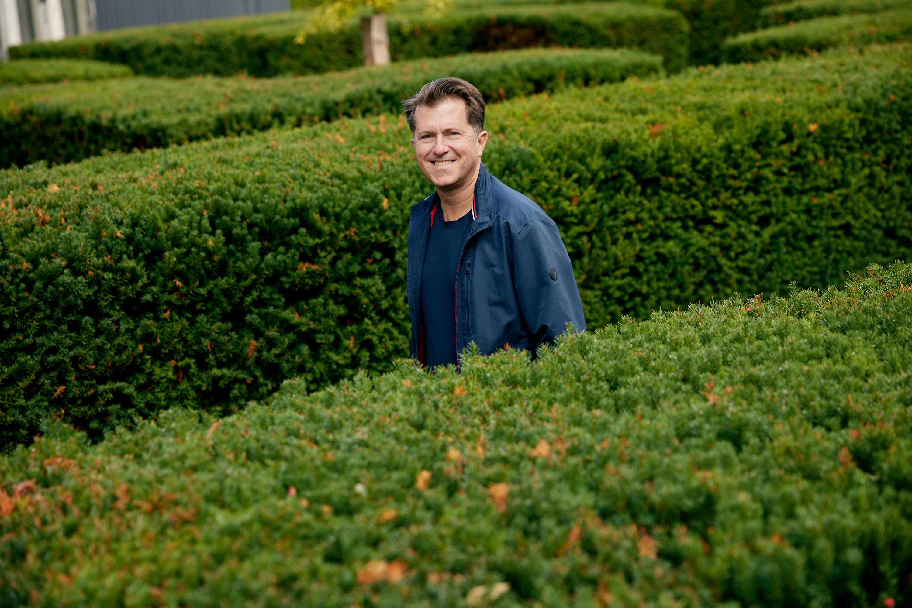 Ein Mann in einer blauen Jacke steht inmitten eines grünen Heckenlabyrinths und lächelt in die Kamera - ein einladendes Geschäftsporträt, das sich perfekt für das Personal Branding eignet. Portrait und Reportage Fotografie für Corporate und Editorial