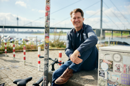 Ein Mann sitzt auf einem Betonblock in der Nähe eines Flusses und lächelt in die Kamera für ein Geschäftsporträt. Im Hintergrund sind eine Brücke und Fahrräder zu sehen sowie Aufkleber auf einem nahe gelegenen Mast und einem Stromkasten. Portrait und Reportage Fotografie für Corporate und Editorial