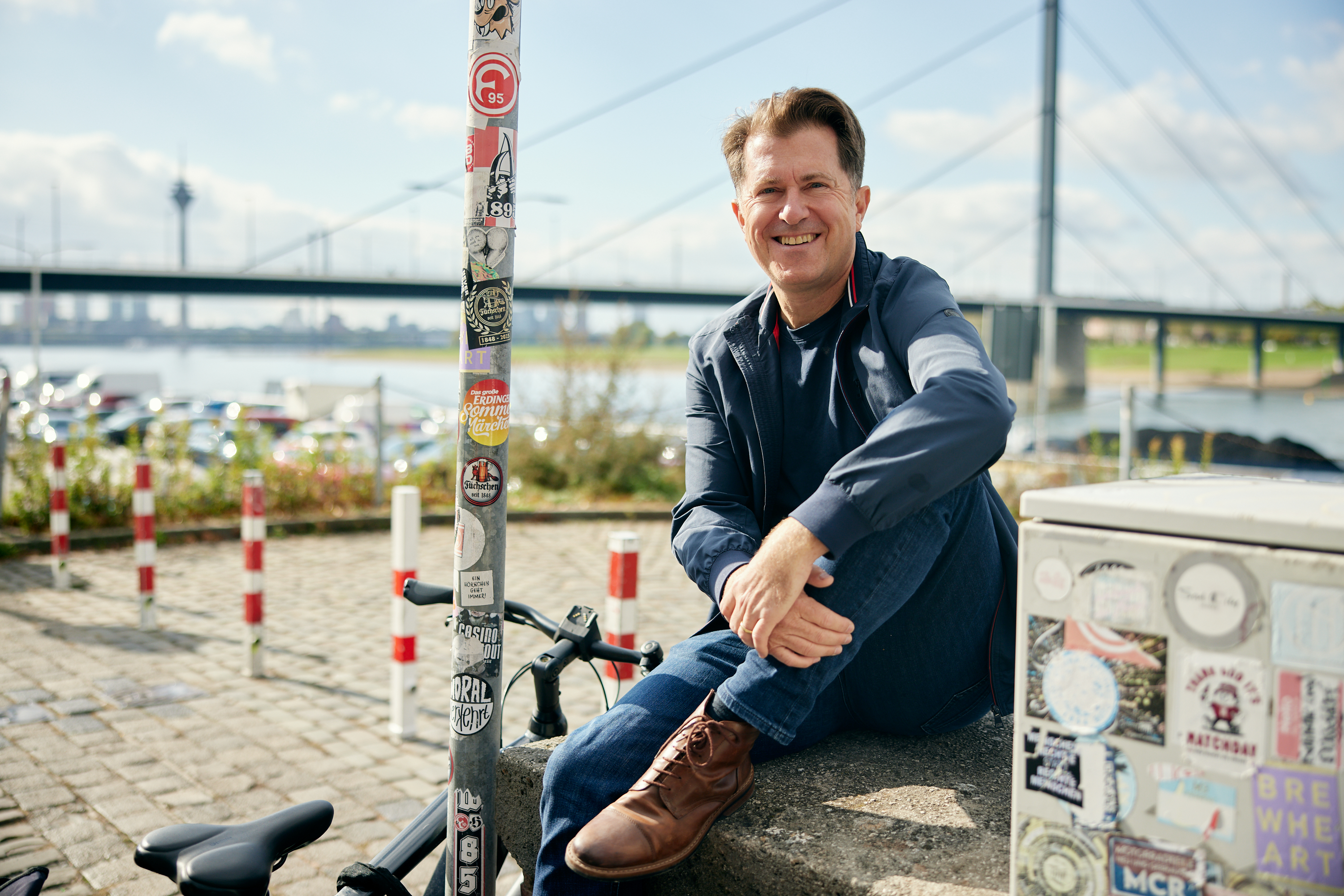 Ein Mann sitzt auf einem Betonblock in der Nähe eines Flusses und lächelt in die Kamera für ein Geschäftsporträt. Im Hintergrund sind eine Brücke und Fahrräder zu sehen sowie Aufkleber auf einem nahe gelegenen Mast und einem Stromkasten. Portrait und Reportage Fotografie für Corporate und Editorial