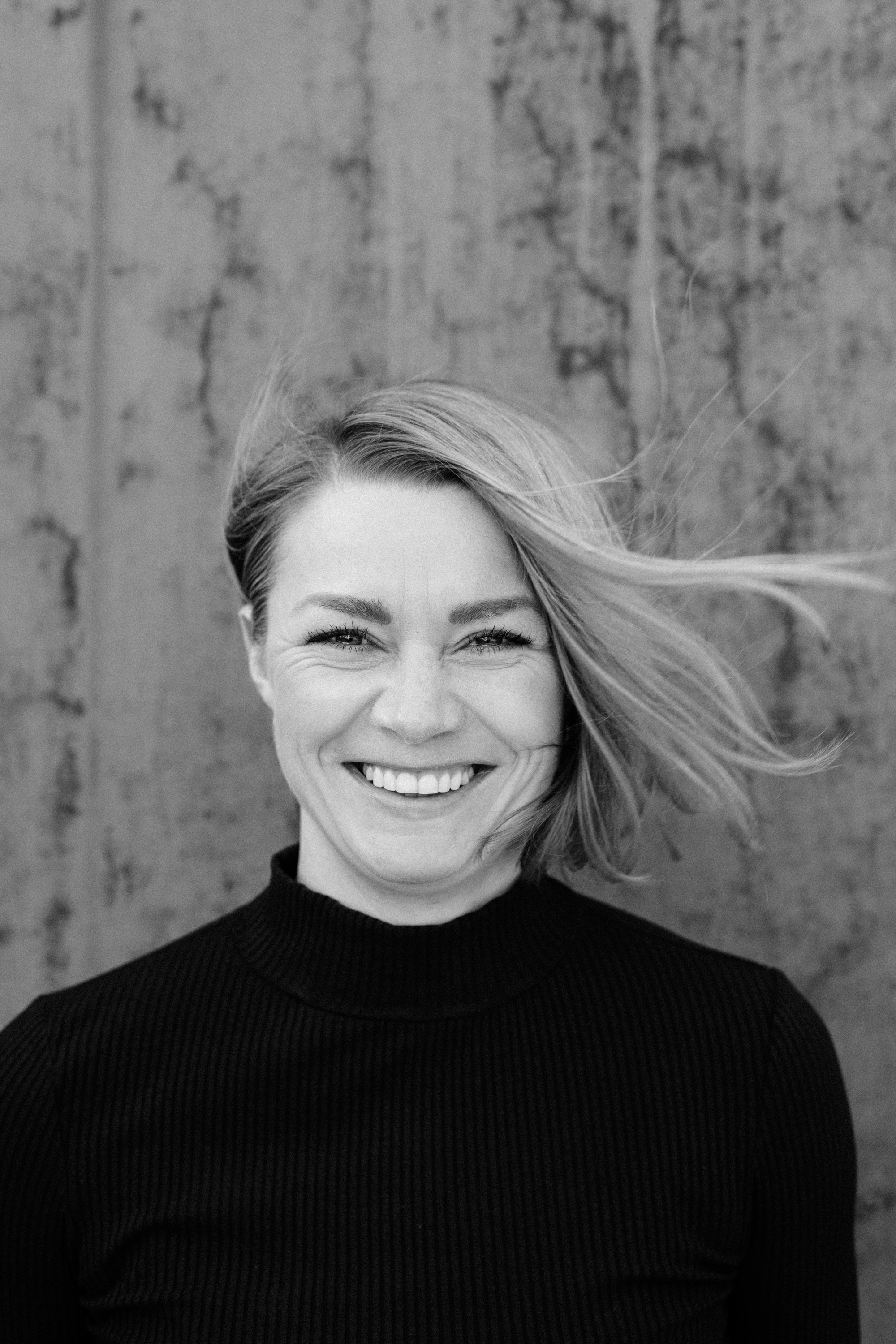 Schwarz-weißes Business-Porträt einer Frau, die vor einer strukturierten Wand steht und selbstbewusst lächelt, während ihr Haar zur Seite geblasen wird - perfekt für Personal Branding oder Unternehmensprofile. Portrait und Reportage Fotografie für Corporate und Editorial