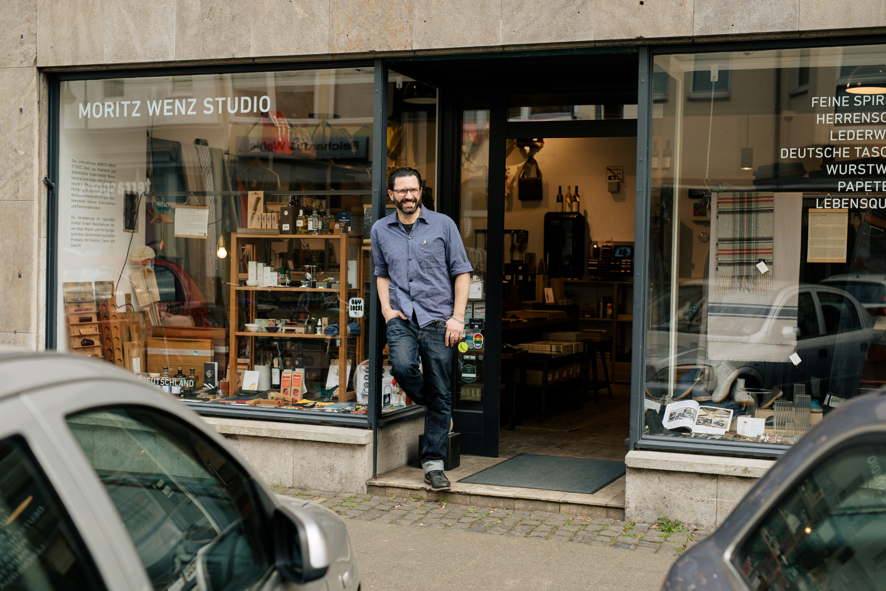 Ein Mann posiert für ein Geschäftsporträt am Eingang des Moritz Wenz Studio, einem Geschäft mit verschiedenen Artikeln im Schaufenster, die sein persönliches Markenzeichen in einer Stadtstraße mit parkenden Autos hervorheben. Portrait und Reportage Fotografie für Corporate und Editorial