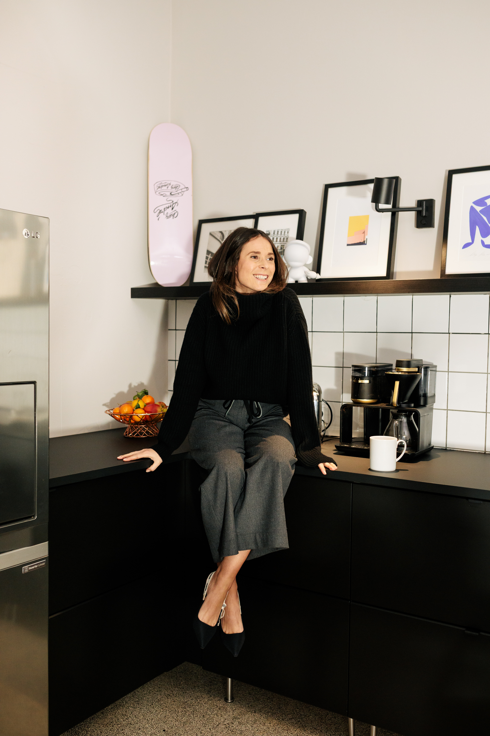 Eine Frau sitzt auf einer schwarzen Küchentheke neben einer Kaffeemaschine, einer Tasse, einer Schale mit Obst und einem gerahmten Kunstwerk. Hinter ihr liegt ein Skateboard auf einem Regal - ein einzigartiges Geschäftsporträt, das modernen Lifestyle mit persönlichem Branding verbindet. Portrait und Reportage Fotografie für Corporate und Editorial
