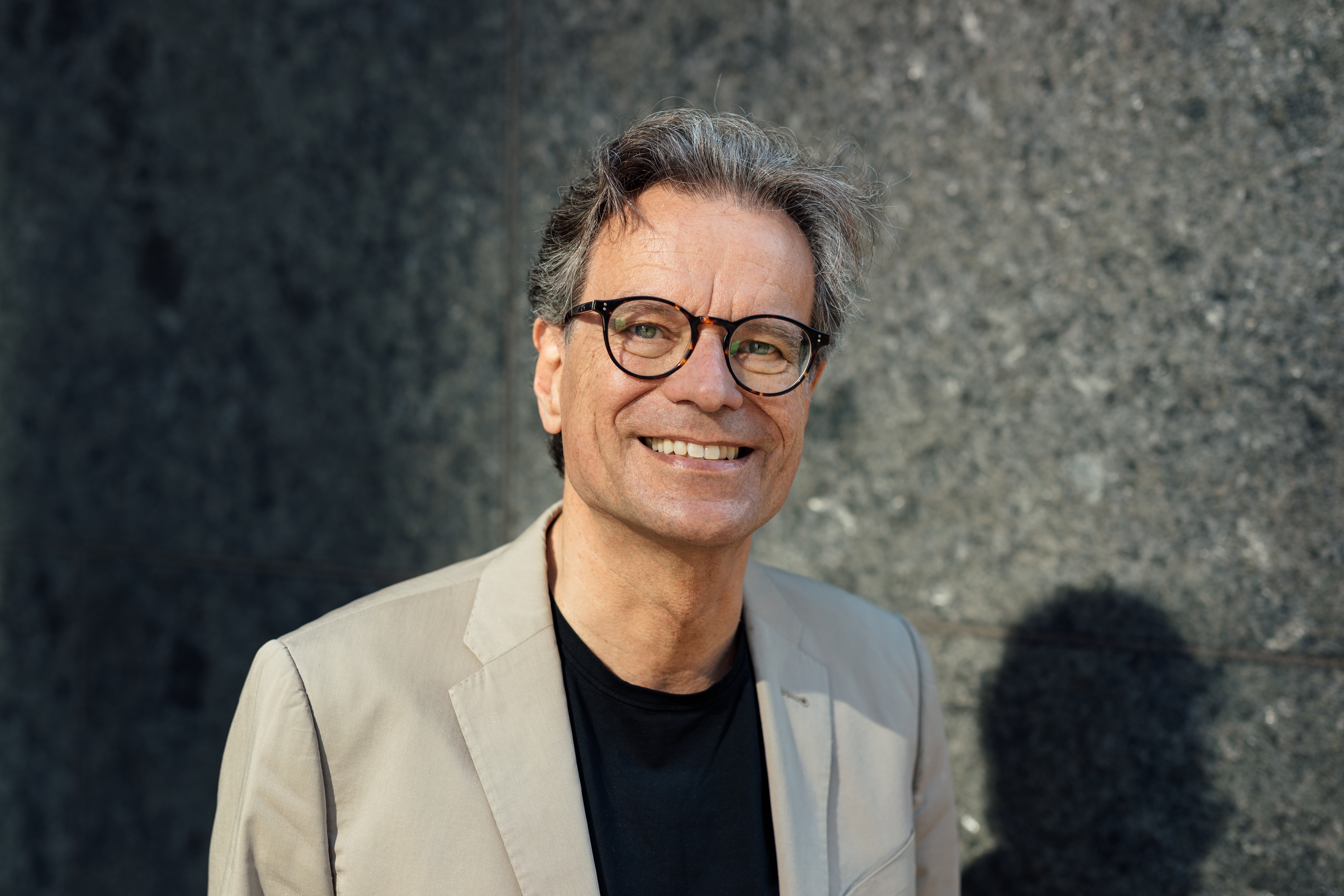 Ein Mann mittleren Alters mit Brille und grauem Haar, der einen beigen Blazer und ein schwarzes Hemd trägt, lächelt vor einer strukturierten grauen Steinwand - ein klassisches Geschäftsporträt, das sich perfekt für die persönliche Markenbildung eignet. Portrait und Reportage Fotografie für Corporate und Editorial