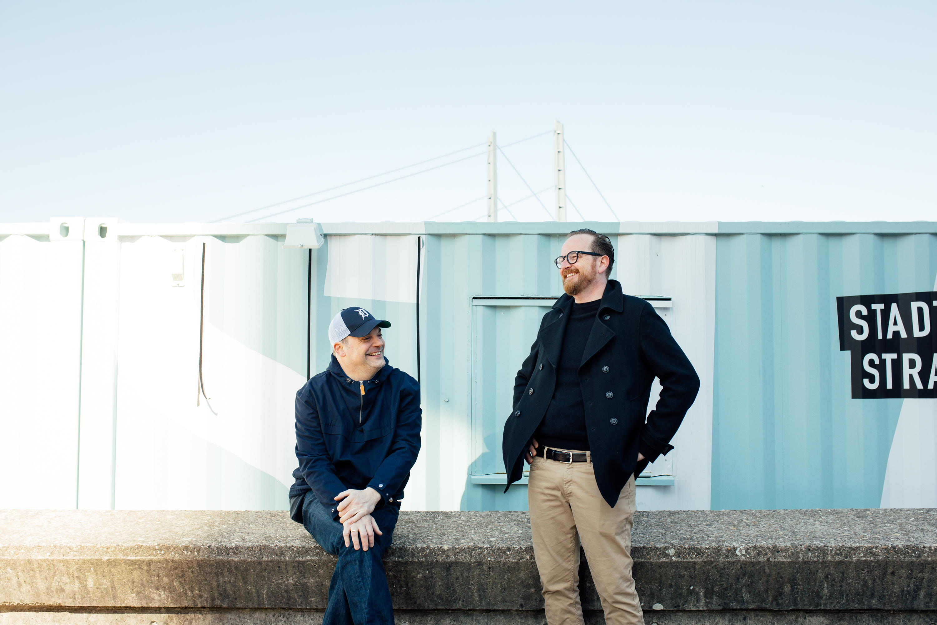 Zwei Männer interagieren draußen vor einem blau-weißen Schiffscontainer; einer sitzt auf einem Sims, während der andere mit den Händen in den Taschen dasteht. So entsteht ein entspanntes und dennoch professionelles Geschäftsporträt, das sich perfekt für das persönliche Branding oder Unternehmensprofile eignet. Portrait und Reportage Fotografie für Corporate und Editorial