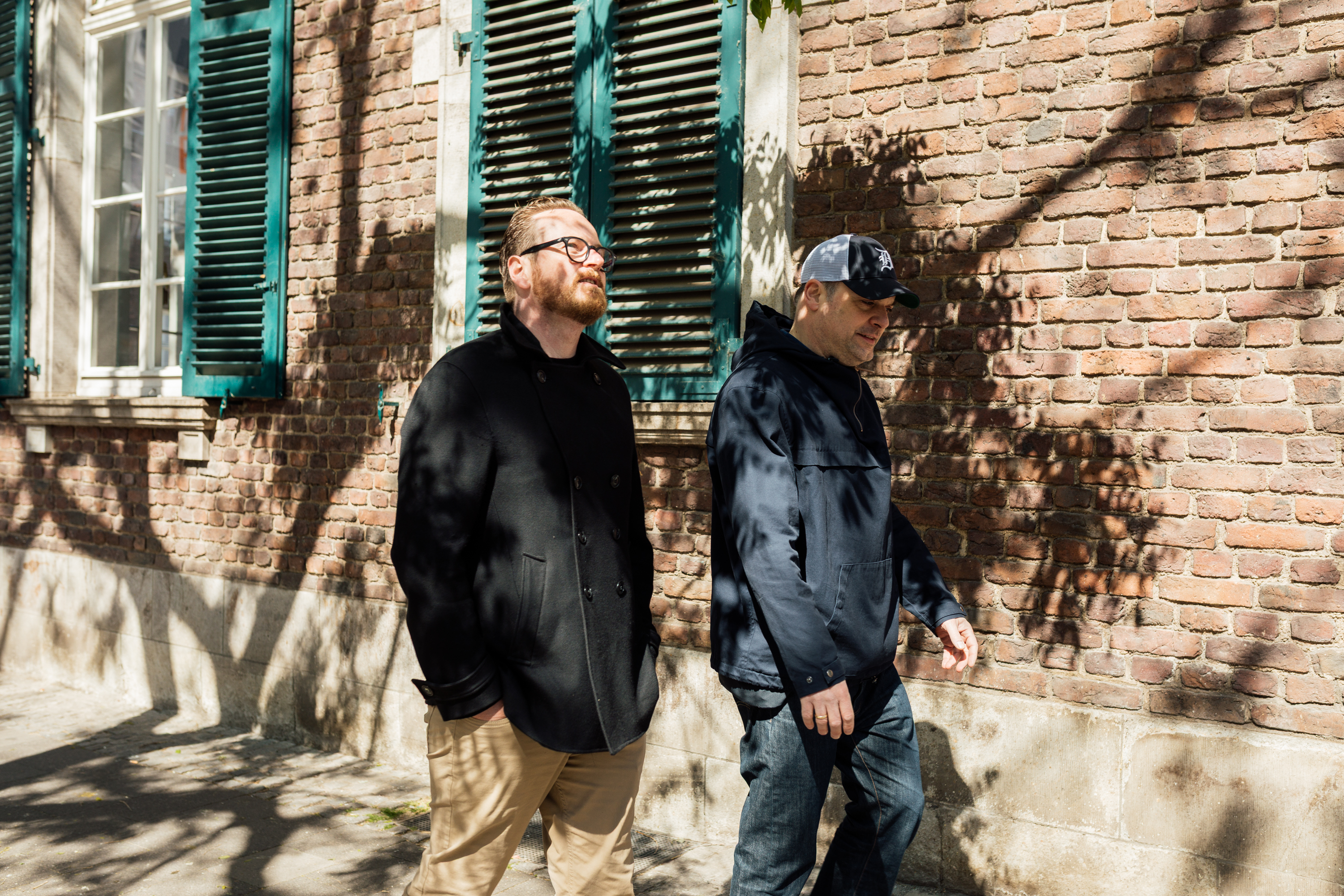 Zwei Männer gehen auf einem sonnenbeschienenen Bürgersteig neben einem Backsteingebäude mit grünen Fensterläden und Baumschatten spazieren - eine perfekte Szene für ein Geschäftsporträt oder ein persönliches Markenfoto. Portrait und Reportage Fotografie für Corporate und Editorial