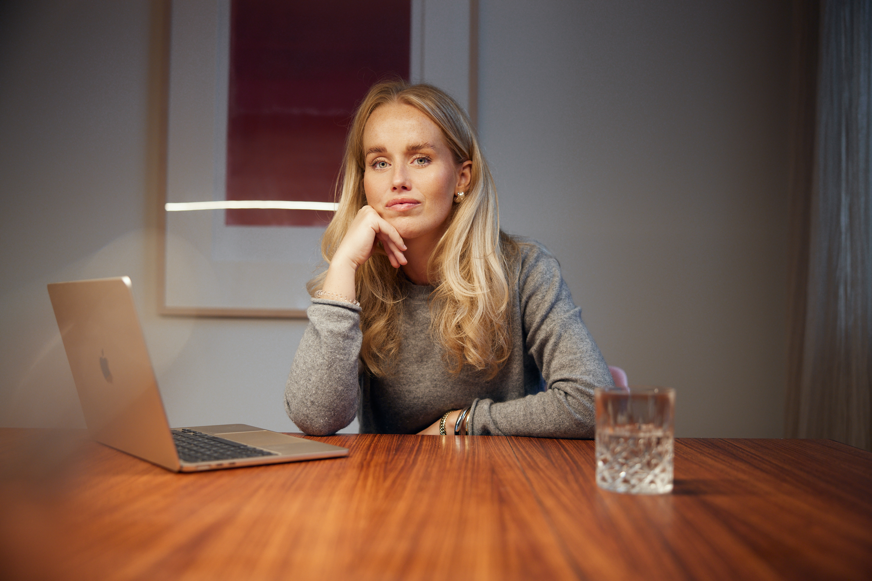 Eine Frau mit langen blonden Haaren sitzt mit einem Laptop und einem Glas Wasser an einem Holztisch und blickt in die Kamera, die Hand auf dem Kinn ruhend - die perfekte Kulisse für ein modernes Business-Porträt oder eine Personal Branding-Session. Portrait und Reportage Fotografie für Corporate und Editorial