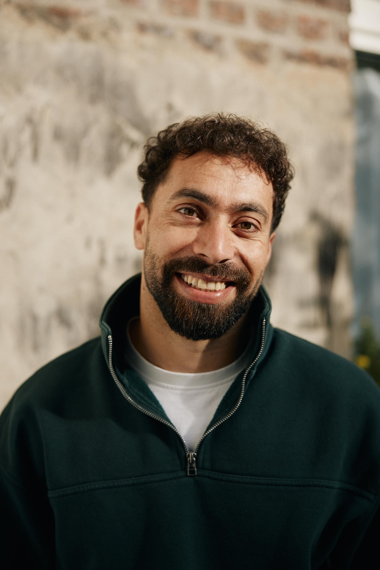 Ein Mann mit lockigem Haar und Bart lächelt warmherzig in einem Leitartikelporträt. Er trägt einen dunklen Pullover mit Reißverschluss über einem weißen Hemd. Er steht vor einer strukturierten Wand mit einem Fenster im Hintergrund und verkörpert den Charme, der jedem Magazinartikel würdig ist. Portrait und Reportage Fotografie für Corporate und Editorial