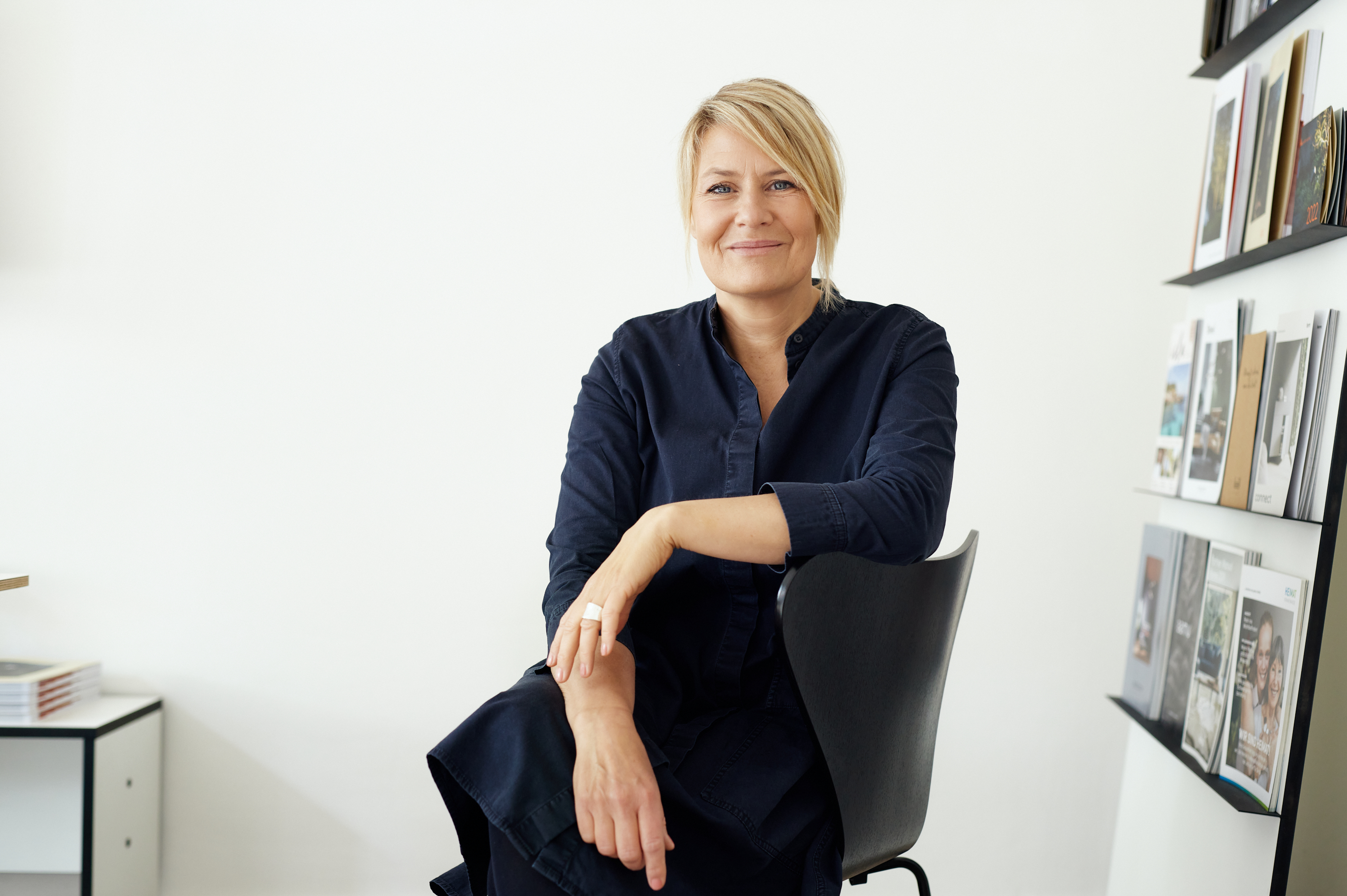 Eine Frau mit blondem Haar in einem marineblauen Kleid sitzt lächelnd auf einem schwarzen Stuhl in einem modernen Raum - perfekt für ein professionelles Geschäftsporträt oder ein persönliches Branding-Foto, mit weißen Wänden und Regalen mit Büchern und Zeitschriften. Portrait und Reportage Fotografie für Corporate und Editorial