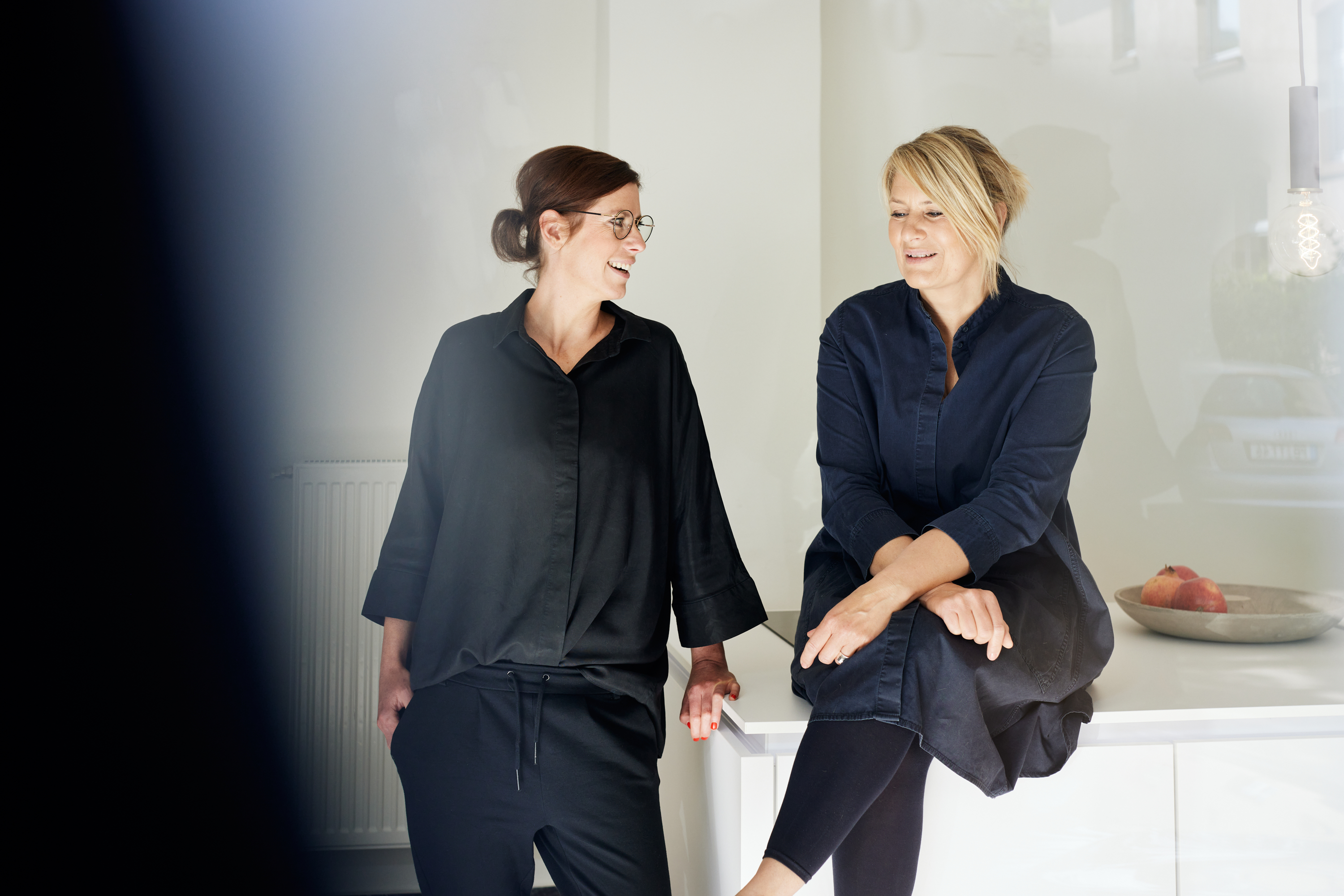 Zwei Frauen in dunkler Kleidung lächeln und unterhalten sich in einer hellen, modernen Küche. Diese entspannte Szene fängt einen persönlichen Branding-Moment ein. Die eine sitzt auf dem Tresen neben einer Schale mit Obst, während die andere in der Nähe steht. Portrait und Reportage Fotografie für Corporate und Editorial