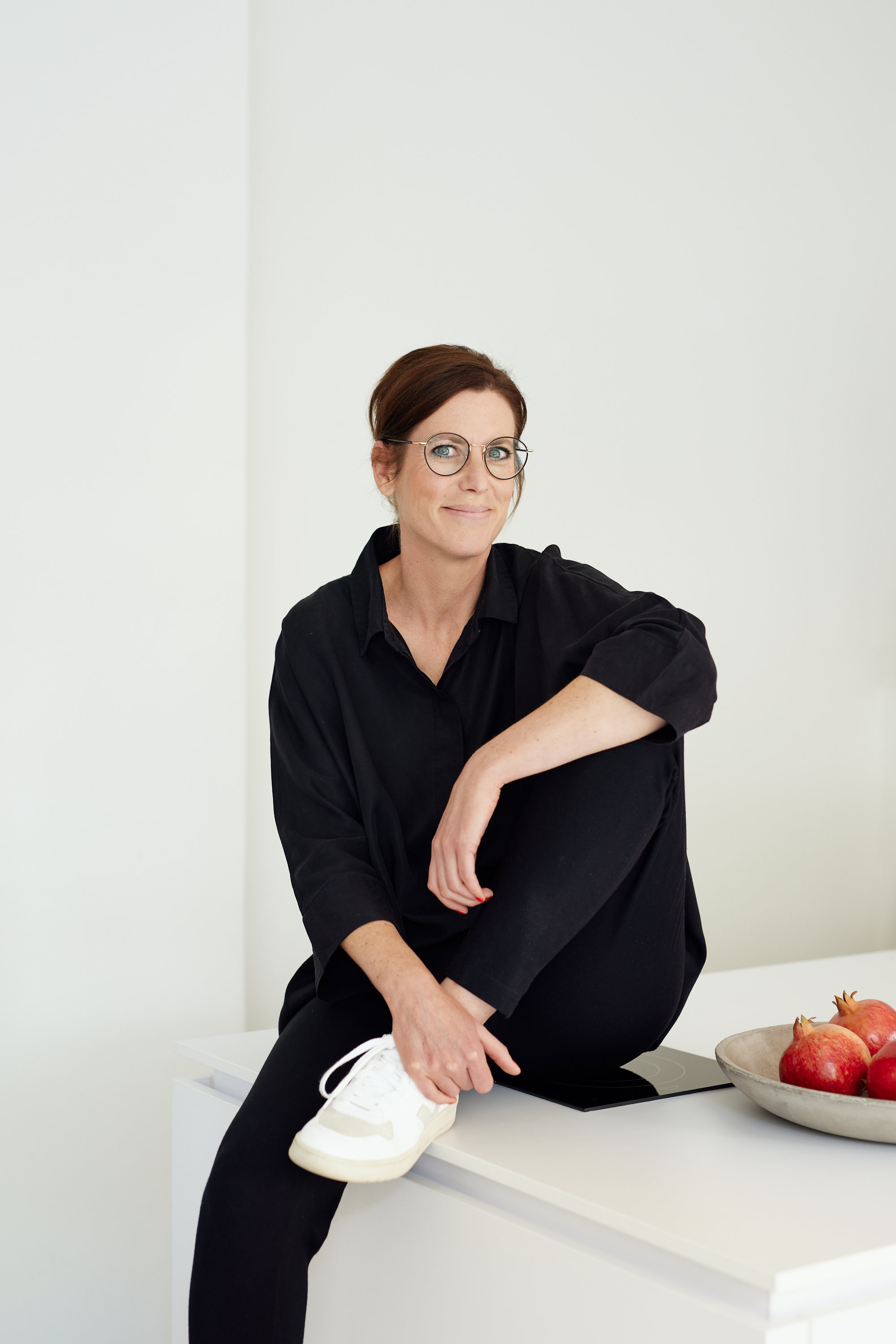 Eine schwarz gekleidete Person mit Brille sitzt auf einer weißen Fläche und hat ein Knie aufgeschlagen - ein modernes Personal Branding-Porträt. Eine Schale mit Granatäpfeln steht auf dem Tresen neben ihr. Portrait und Reportage Fotografie für Corporate und Editorial