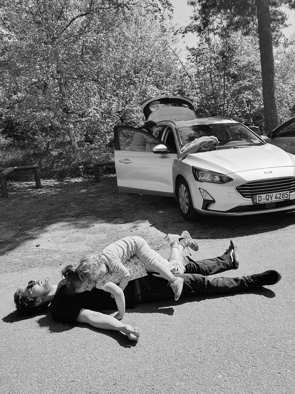 Zwei Menschen liegen auf dem Bürgersteig vor einem geparkten weißen Auto mit offenem Kofferraum, umgeben von Bäumen, und schaffen eine heitere Szene, die an einen Sommertag in Düsseldorf, NRW, erinnert. Portrait und Reportage Fotografie für Corporate und Editorial