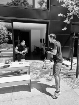 Auf einer schicken Düsseldorfer Terrasse entspannen sich zwei Männer – einer hält den Moment mit seiner Kamera fest, während der andere in ein spannendes Buch vertieft ist. In der Nähe steht ein authentischer Esstisch, der darauf wartet, ein köstliches Essen serviert zu bekommen. Portrait und Reportage Fotografie für Corporate und Editorial