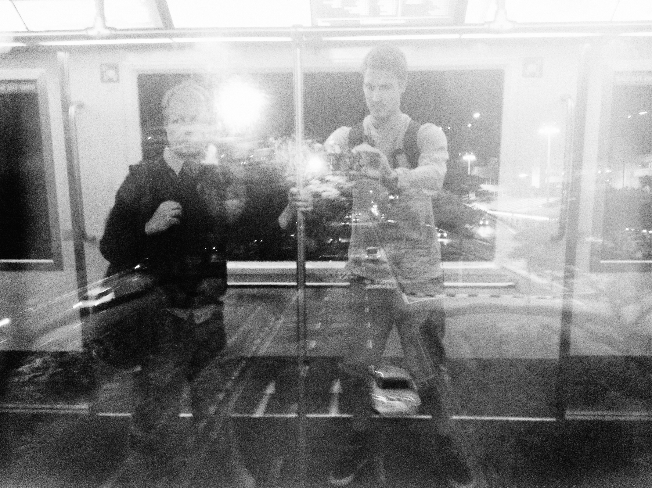 Zwei Personen fotografieren ein authentisches Foto in einer reflektierenden Glasscheibe. Durch die Spiegelung erwacht die nächtliche Stadtszene NRWs mit funkelnden Lichtern zum Leben. Portrait und Reportage Fotografie für Corporate und Editorial