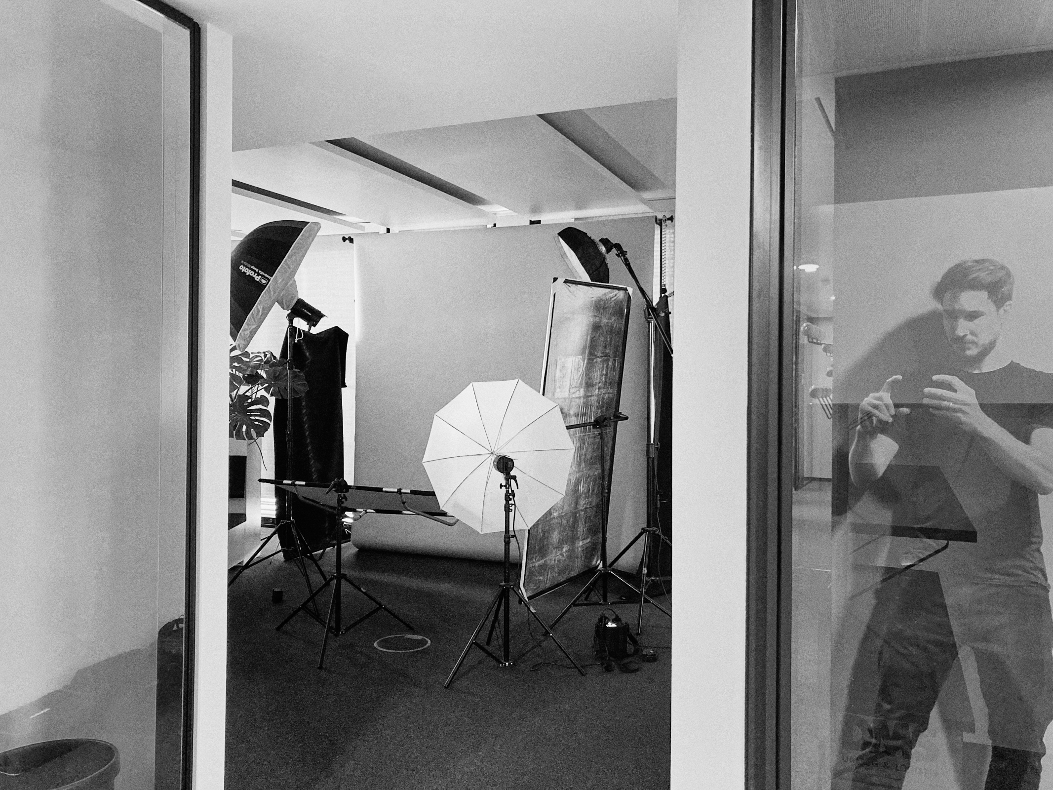 In einem Düsseldorfer Fotostudio wird Beleuchtungsausrüstung wie Schirme und Softboxen vor einem Hintergrund aufgebaut. In der Spiegelung rechts ist eine Person zu sehen, die wahrscheinlich einen Moment mit ihrem Telefon festhält. Perfekt für jedes Unternehmen, das professionelle Bilder sucht. Portrait und Reportage Fotografie für Corporate und Editorial