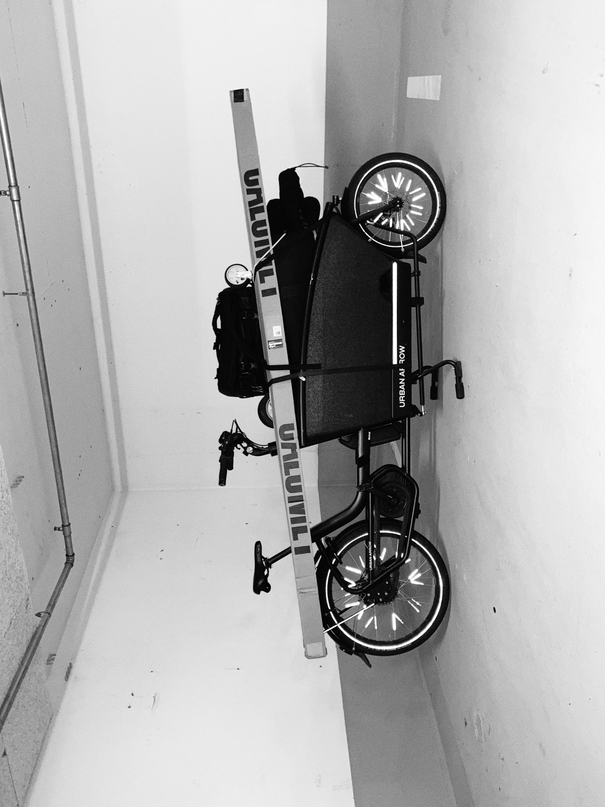 Ein umgedrehtes Bild eines Lastenfahrrads mit dem „Urban Arrow“-Logo und großem Laderaum, das in der monochromen Umgebung Düsseldorfs an einer Wand geparkt ist. Portrait und Reportage Fotografie für Corporate und Editorial