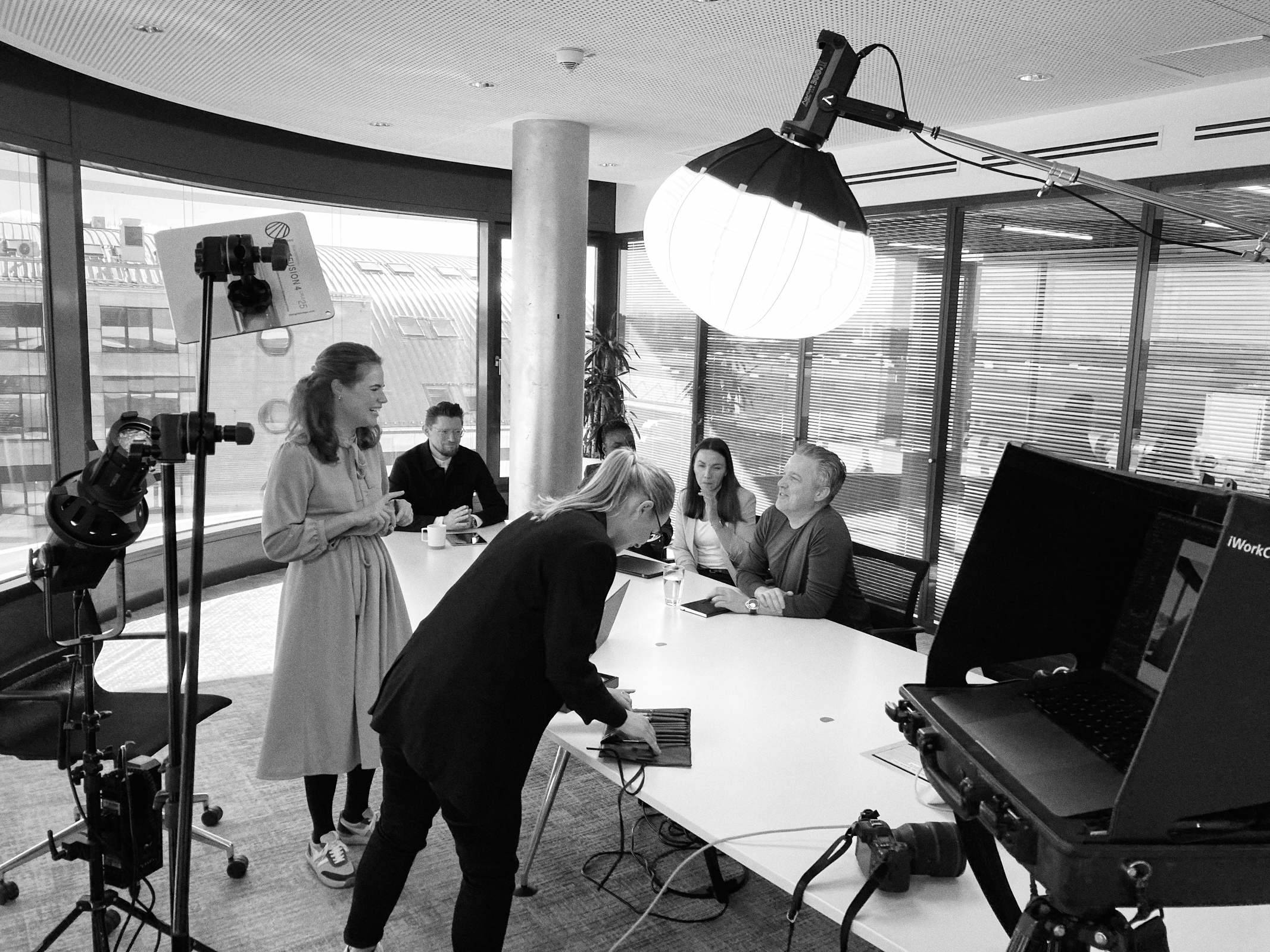 Menschen sitzen um einen Konferenztisch in einem modernen Büro in NRW und diskutieren authentisch. Eine Frau steht daneben, während eine andere Person die Geräte einrichtet. Eine große Leuchte sorgt für ausreichend Licht für die Versammlung, als ob sie von einem erfahrenen Fotografen eingefangen worden wäre. Portrait und Reportage Fotografie für Corporate und Editorial