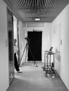 Eine Person steht in einem Düsseldorfer Hauseingang und blickt in einen kleinen Raum, in dem ein echter Fotograf eine Kamera auf einem Stativ, einen schwarzen Hintergrund und einen Wagen mit einem Laptop aufgestellt hat. Die Decke ist mit Holzlamellen verkleidet, was der professionellen Umgebung Wärme verleiht. Portrait und Reportage Fotografie für Corporate und Editorial