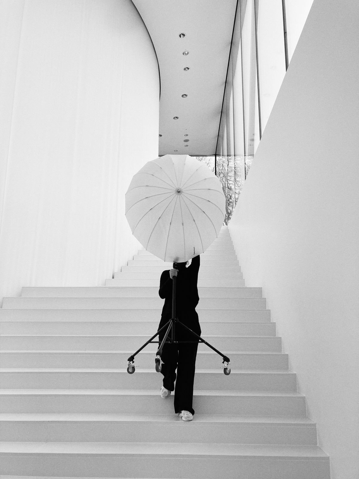 Eine Person trägt einen großen Regenschirm auf Rollen eine moderne, minimalistische Treppe in Düsseldorf hinauf. Der Raum zeichnet sich durch hohe Decken und klare Linien aus – eine wahrhaft authentische Mischung aus Stil und Funktion. Portrait und Reportage Fotografie für Corporate und Editorial