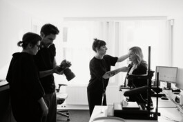 In einem Düsseldorfer Büro arbeitet ein Team aus vier Personen zusammen. Eine Person frisiert die Haare einer anderen, während ein Fotograf mit der Kamera hantiert. Auf den Schreibtischen stehen Computer, die die kreative Energie einfangen, die NRW so einzigartig macht. Portrait und Reportage Fotografie für Corporate und Editorial