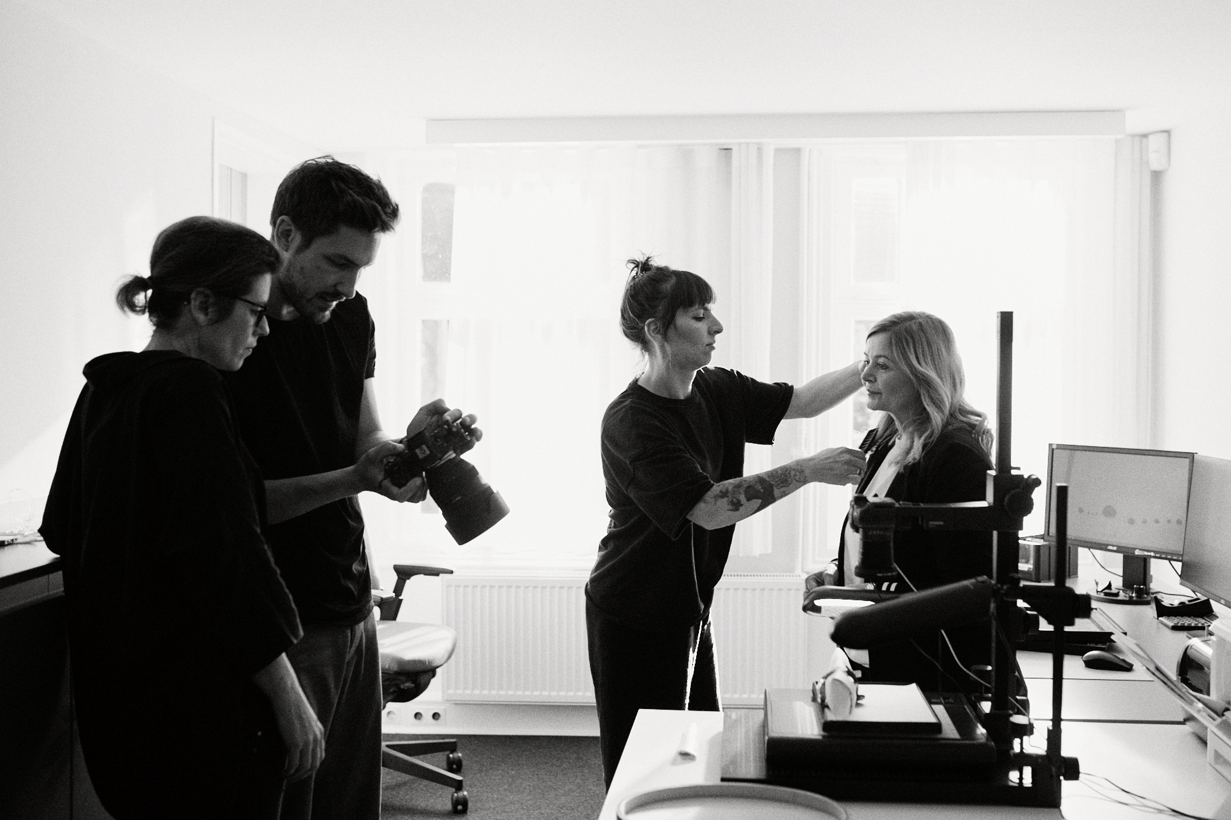 In einem Düsseldorfer Büro arbeitet ein Team aus vier Personen zusammen. Eine Person frisiert die Haare einer anderen, während ein Fotograf mit der Kamera hantiert. Auf den Schreibtischen stehen Computer, die die kreative Energie einfangen, die NRW so einzigartig macht. Portrait und Reportage Fotografie für Corporate und Editorial