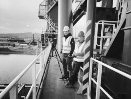 Zwei Arbeiter in Schutzausrüstung stehen authentisch auf einer Industriebühne in der Nähe von Geländer und Anlagen, im Hintergrund sind die Düsseldorfer Industriebauten und ein Gewässer zu sehen. Portrait und Reportage Fotografie für Corporate und Editorial