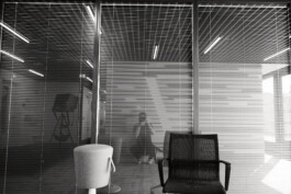 Schwarz-Weiß-Bild eines leeren Raums mit Glaswänden, Jalousien und einem Stuhl. Spiegelungen im Glas lassen subtil eine sitzende Person erkennen. Die Kulisse ruft eine authentische Atmosphäre hervor, die an ein elegantes Düsseldorfer Büro erinnert. Portrait und Reportage Fotografie für Corporate und Editorial