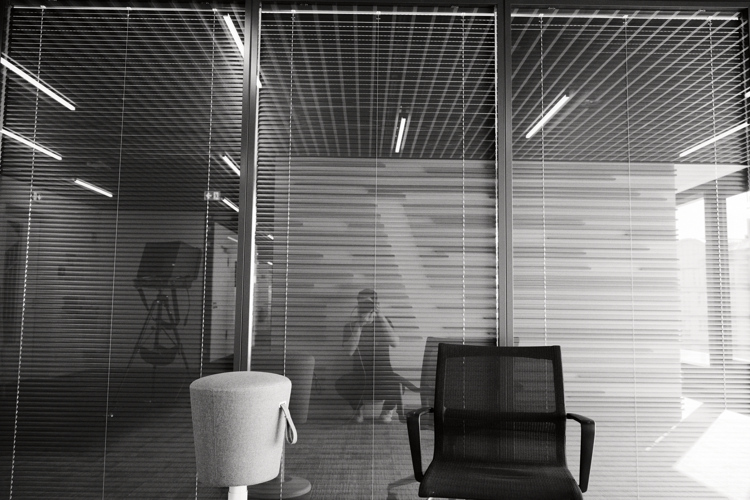 Schwarz-Weiß-Bild eines leeren Raums mit Glaswänden, Jalousien und einem Stuhl. Spiegelungen im Glas lassen subtil eine sitzende Person erkennen. Die Kulisse ruft eine authentische Atmosphäre hervor, die an ein elegantes Düsseldorfer Büro erinnert. Portrait und Reportage Fotografie für Corporate und Editorial