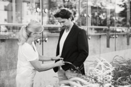 In der ungezwungenen Atmosphäre Düsseldorfs richtet eine Frau vor dem Hintergrund glatter Glasscheiben aufmerksam die Jacke eines Mannes, als ob sie von einem geschickten Fotografen eingefangen worden wäre. Portrait und Reportage Fotografie für Corporate und Editorial