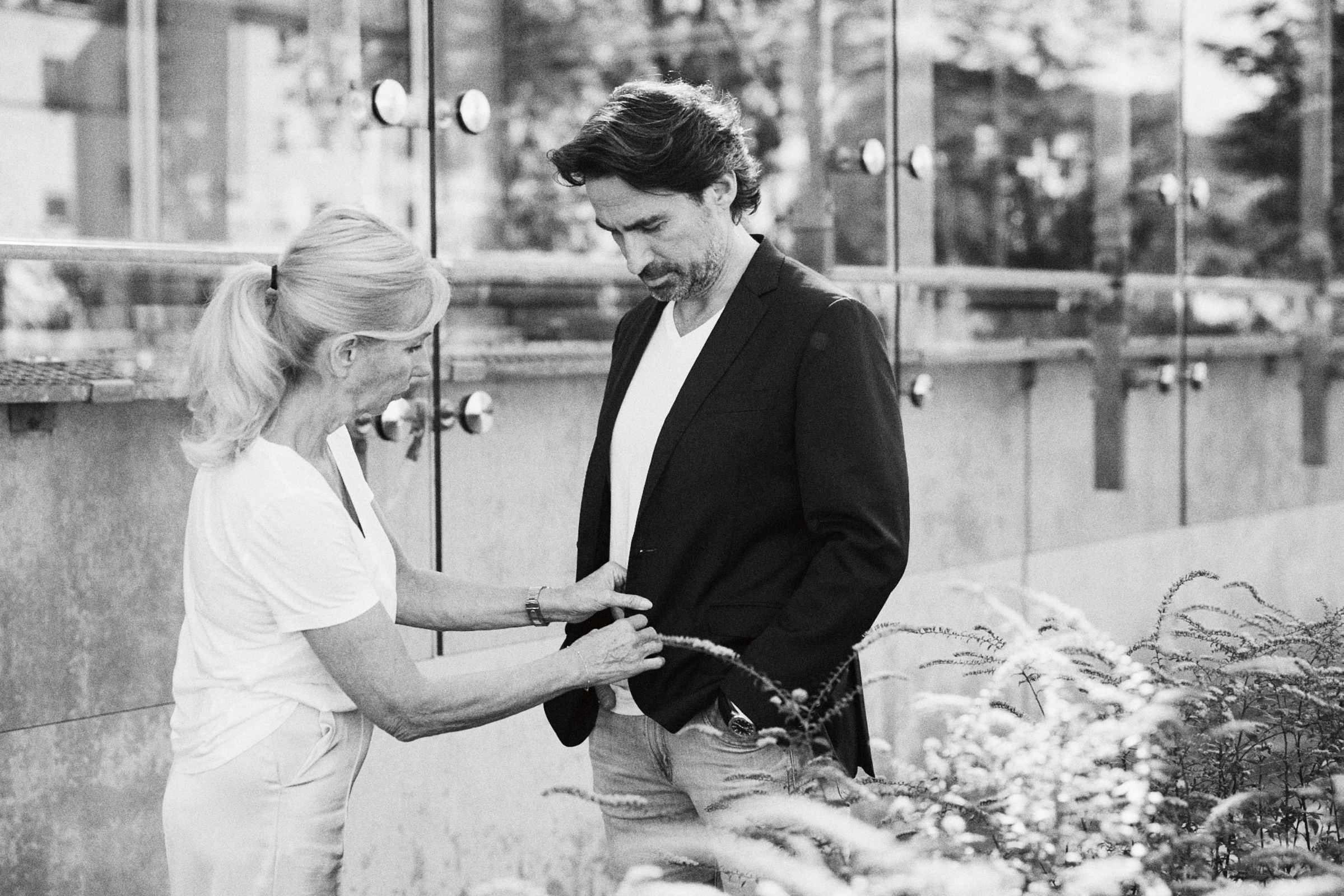 In der ungezwungenen Atmosphäre Düsseldorfs richtet eine Frau vor dem Hintergrund glatter Glasscheiben aufmerksam die Jacke eines Mannes, als ob sie von einem geschickten Fotografen eingefangen worden wäre. Portrait und Reportage Fotografie für Corporate und Editorial