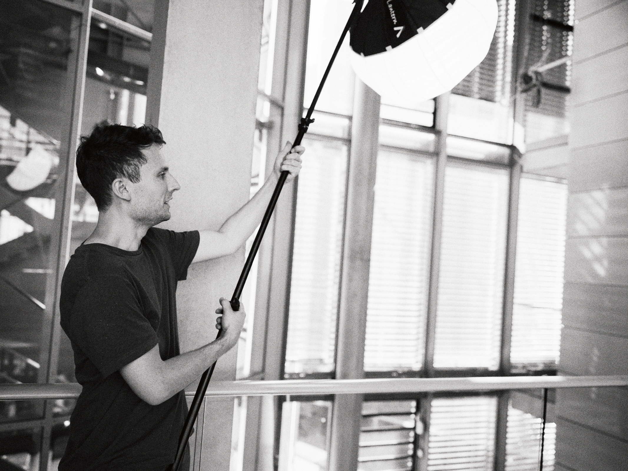 Ein Fotograf im T-Shirt richtet eine große Deckenleuchte auf einem Stativ aus und perfektioniert die Szene in einem Düsseldorfer Innenraum, der mit glatten Glas- und Metallelementen geschmückt ist und die Essenz des modernen Unternehmensdesigns einfängt. Portrait und Reportage Fotografie für Corporate und Editorial