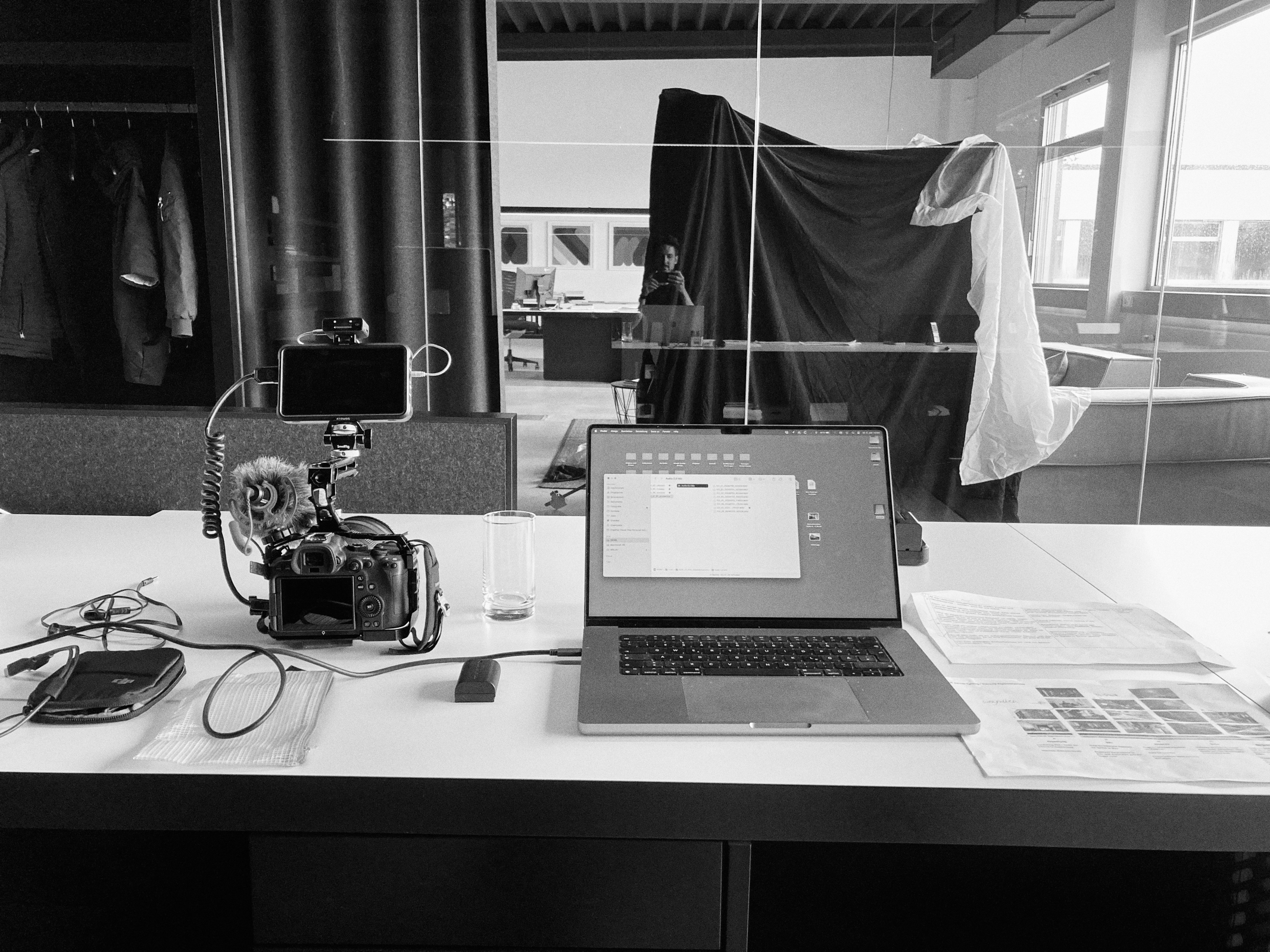 Ein Schwarzweißfoto zeigt eine authentische Schreibtischeinrichtung in Düsseldorf mit Laptop, Kamera auf Stativ, Wasserglas, Papieren und Kabeln. Im Fenster hinter dem Schreibtisch ist die Spiegelung eines Fotografen zu sehen. Portrait und Reportage Fotografie für Corporate und Editorial