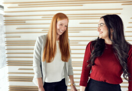 Zwei Frauen stehen vor einer modernen Wand mit Holzakzenten in Aachen und lächeln sich herzlich an. Ihr lebhafter Austausch verkörpert die Essenz eines starken Employer Brandings und zeigt eine Unternehmenskultur, in der Zusammenarbeit und positives Miteinander gedeihen. Portrait und Reportage Fotografie für Corporate und Editorial