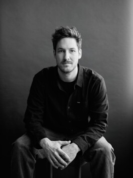 Eine Person sitzt auf einem Hocker vor einem dunklen Hintergrund, trägt ein dunkles Hemd und Jeans und blickt mit einer Intensität in die Kamera, die die Essenz der Porträtfotografie einfängt. Dieser fesselnde Moment wurde von einem talentierten Fotografen in Düsseldorf gekonnt festgehalten. Portrait und Reportage Fotografie für Corporate und Editorial